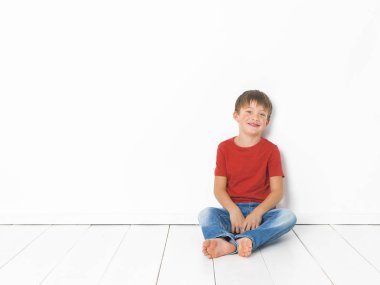 kırmızı t-shirt ve mavi kot beyaz arka plan önünde beyaz ahşap zemin üzerinde oturan sevimli sarışın çocuk