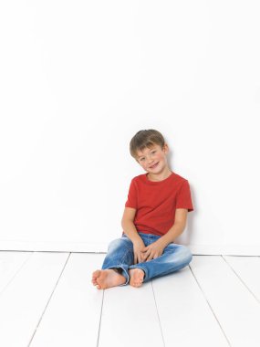 kırmızı t-shirt ve mavi kot beyaz arka plan önünde beyaz ahşap zemin üzerinde oturan sevimli sarışın çocuk