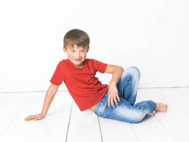 kırmızı t-shirt ve mavi kot beyaz arka plan önünde beyaz ahşap zemin üzerinde oturan sevimli sarışın çocuk