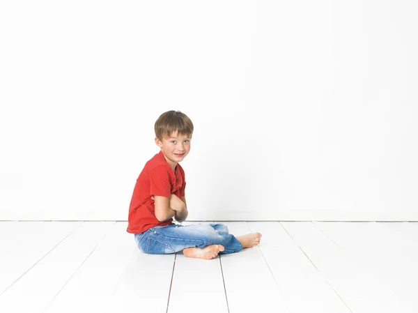 可爱的金发男孩在红色T恤和蓝色牛仔裤坐在白色木地板上 白色背景前交叉的手臂 — 图库照片