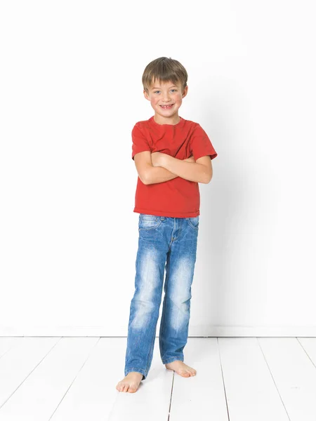 可爱的金发男孩在红色T恤和蓝色牛仔裤站在白色木地板上交叉的手臂在白色背景前 — 图库照片