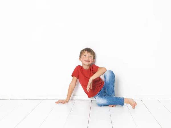 可爱的金发男孩在红色T恤和蓝色牛仔裤坐在白色木地板前的白色背景 — 图库照片