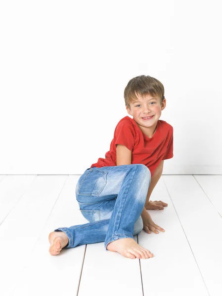 白い背景の前に白い木製の床に座っている赤いTシャツと青いジーンズのかわいいブロンドの男の子 — ストック写真