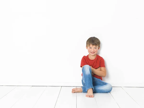 可爱的金发男孩在红色T恤和蓝色牛仔裤坐在白色木地板上 白色背景前交叉的手臂 — 图库照片