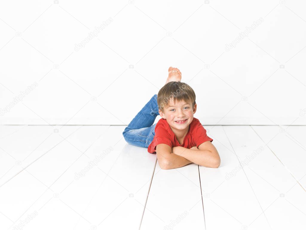 cute blond boy in red t-shirt and blue jeans lying with crossed arms on white wooden floor in front of white background