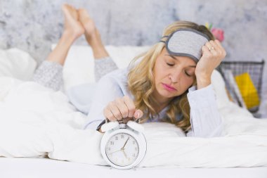 blonde pretty young woman wearing pajamas lying in bed with ringing alarm clock at home in early morning  clipart