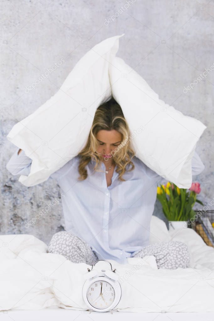 blonde young woman wearing pajamas closing ears with pillows while sitting in bed with ringing alarm clock at home in early morning 