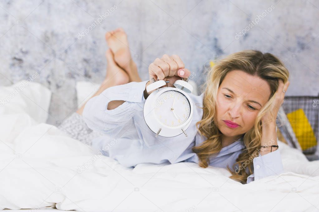 blonde pretty young woman wearing pajamas lying in bed with ringing alarm clock at home in early morning 