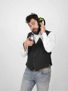 siyah sakallı komik adam kulaklık müzik dinlerken ve beyaz stüdyo duvar önünde kamera işaret ederken akıllı telefon tutarak