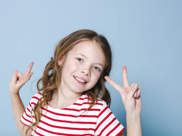 pretty girl with curly hair in striped t-shirt showing peace signs and looking at camera in front of blue background