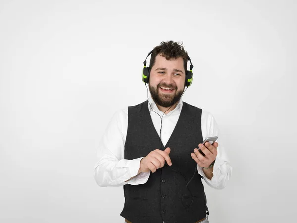 smiling man with black beard listening to music in headphones and holding smartphone while looking at down in front of white studio wall