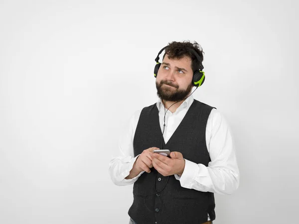pensive man with black beard listening to music in headphones and holding smartphone while looking up in front of white studio wall