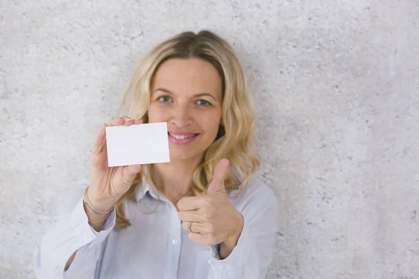 happy beautiful blonde woman holding empty white card in front of concrete background