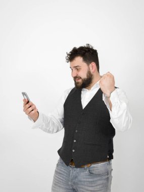 Siyah sakallı genç adam akıllı telefon tutuyor ve kollarını kaldırmış beyaz arka planda zafer kutlamaları yapıyor.