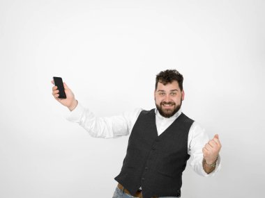 Siyah sakallı genç adam akıllı telefon tutuyor ve kollarını kaldırmış beyaz arka planda zafer kutlamaları yapıyor.