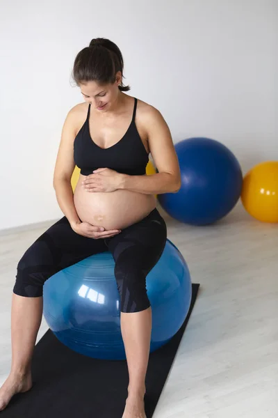 美丽的年轻孕妇穿着黑色运动服轻轻地拥抱肚子 而坐在健身球和放松后健身锻炼在轻工作室 — 图库照片