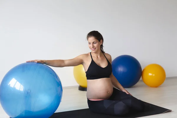 マットの上に座って 軽いスタジオで有酸素ボールに傾いている間 フィットネス運動の後にリラックスした黒のスポーツウェアの美しい若い妊娠中の女性 — ストック写真