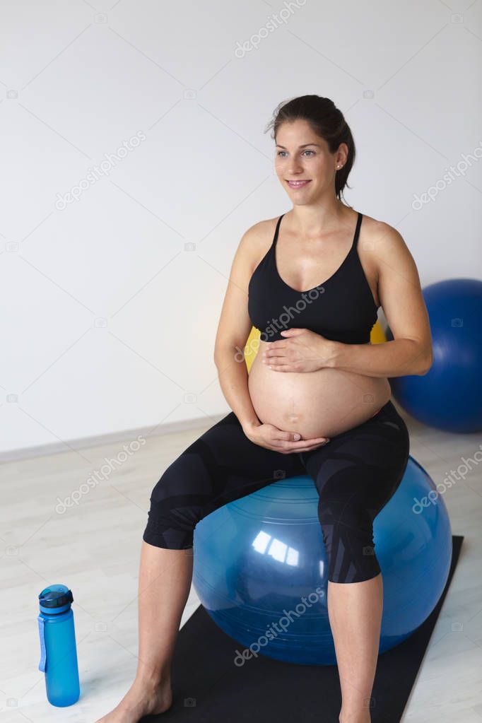 beautiful young pregnant woman in black sportswear gently hugging belly with hands while sitting on fitness ball and relaxing after fitness exercise in light studio