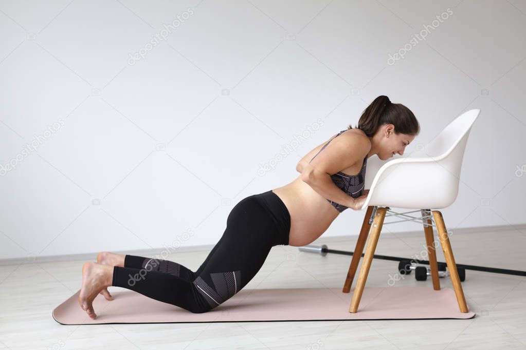 beautiful young pregnant woman in black sportswear doing chair push-up exercise in light studio