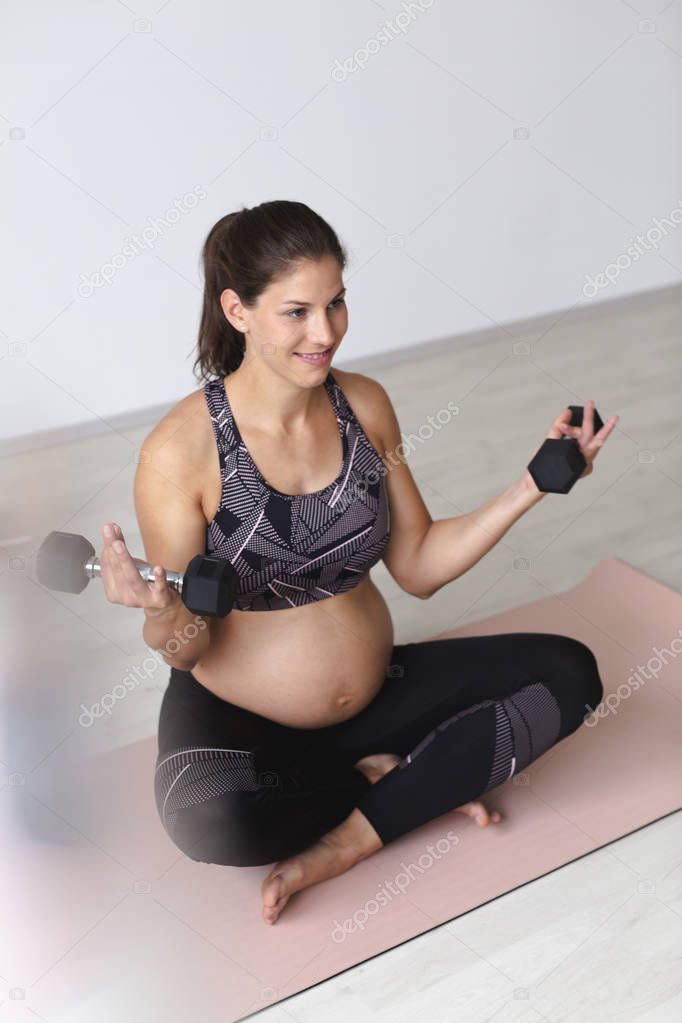 beautiful young pregnant woman in black sportswear holding dumbbells in hands and sitting in easy pose on mat in light studio 