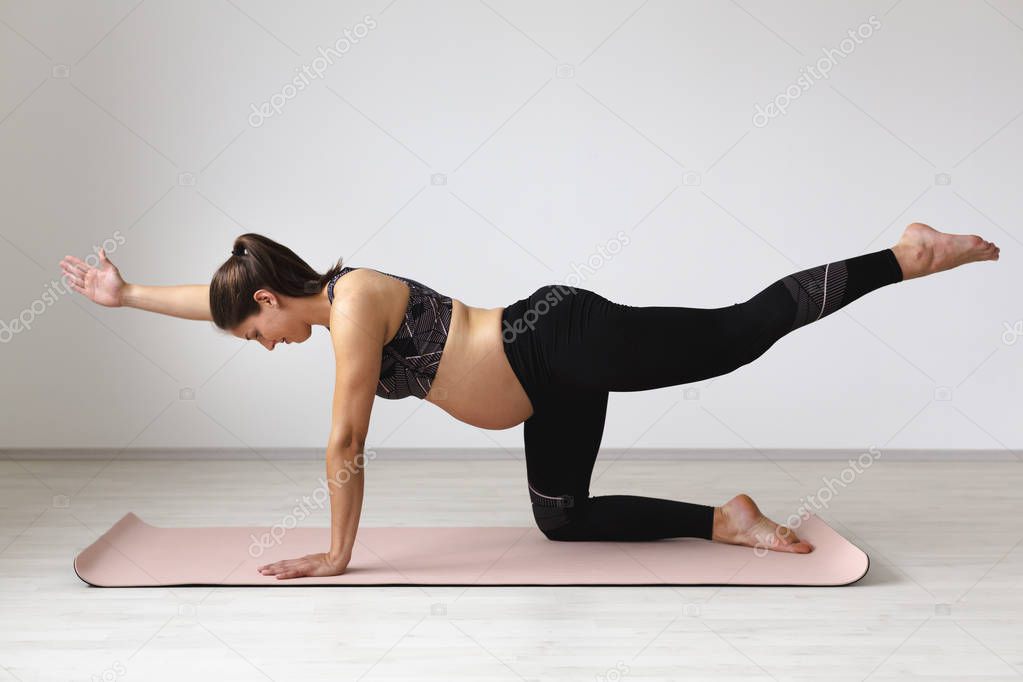 pregnant sporty yogi woman in black sportswear stretching on mat while standing on knee in light studio
