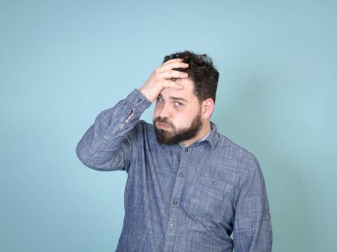 Kaş çatan, siyah saçlı, siyah sakallı, acı dolu başını kaldırıp mavi duvarın arkasında poz veren bir adam.