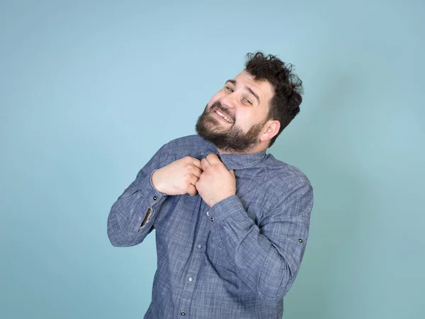 man with black hair and black beard fastening shirt while posing in front of blue wall background