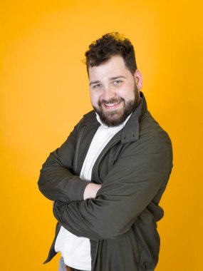 Siyah sakallı, siyah saçlı, beyaz gömlekli, siyah ceketli mutlu bir adam kameraya bakıyor ve stüdyo turuncu arka planında çapraz kollarla poz verirken gülümsüyor. 