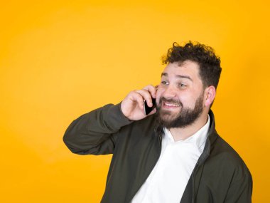 Mutlu siyah sakallı adam turuncu arka planda poz verirken gülümsüyor ve akıllı telefonla konuşuyor. 