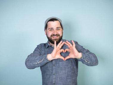 Gri şapkalı ve kot pantolonlu genç bir adam kalp şeklini, aşk hareketini ve mavi arka planda gülümsemesini gösteriyor. 