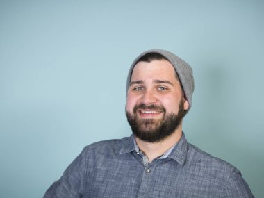 Gri şapkalı ve kot gömlekli genç bir adam gülümsüyor ve mavi arka plandaki kameraya bakıyor. 