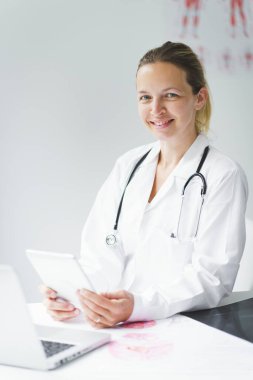 Genç, sarışın doktor muayenehanesinde oturuyor, tableti üzerinde çalışıyor ve sınav sonuçlarına bakıyor.