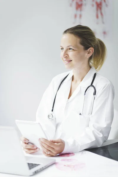 young, blonde doctor is sitting in her practice, working on her tablet and looking at examination results