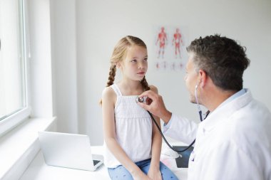 Beyaz önlüklü erkek doktor muayenehanesinde genç ve güzel bir kızı muayene ediyor.