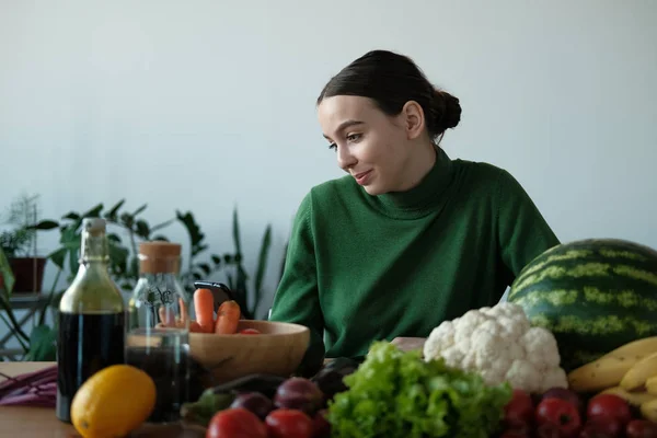 young vegan woman using phone at kitchen table with veggies in home interior