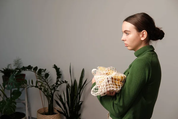young woman holding string bag with groceries in home interior