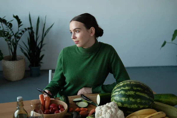 young vegan woman using phone at kitchen table with veggies in home interior