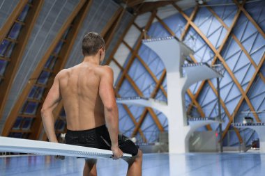 kapalı yüzme havuzunda sıçrama tahtası üzerinde oturan genç erkek yüzücü