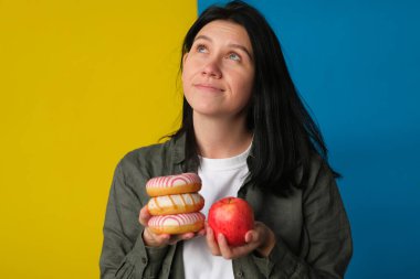 Elinde donut ve mavi ve sarı arka planda taze kırmızı elma tutan kararsız bir kız.