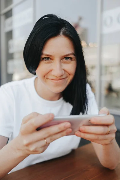 Młoda kobieta z czarnymi włosami w białej koszulce siedzi na otwartej werandzie w kawiarni i używa smartfona czekając na zamówienie i jedzenie na wynos.. — Zdjęcie stockowe