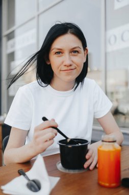 Beyaz tişörtlü, siyah saçlı genç bir kadın bir kafedeki açık bir verandada oturup tek kullanımlık plastik tabaklardan yemek yiyor.. 