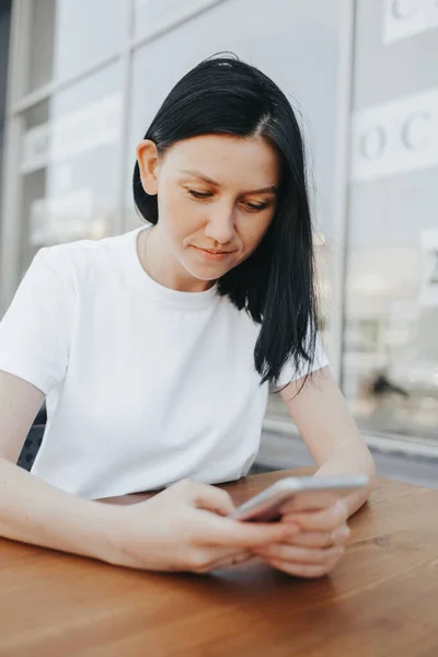 Молодая женщина с черными волосами в белой футболке сидит на открытой веранде в кафе и использует смартфон, ожидая свой заказ и еду на вынос . — стоковое фото