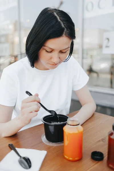 Una giovane donna con i capelli neri in una maglietta bianca siede su una veranda aperta in un caffè e mangia cibo da piatti di plastica usa e getta.. — Foto Stock