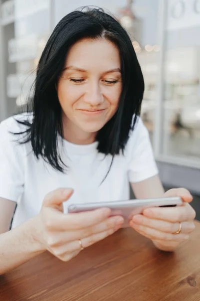 Молодая женщина с черными волосами в белой футболке сидит на открытой веранде в кафе и использует смартфон, ожидая свой заказ и еду на вынос . — стоковое фото