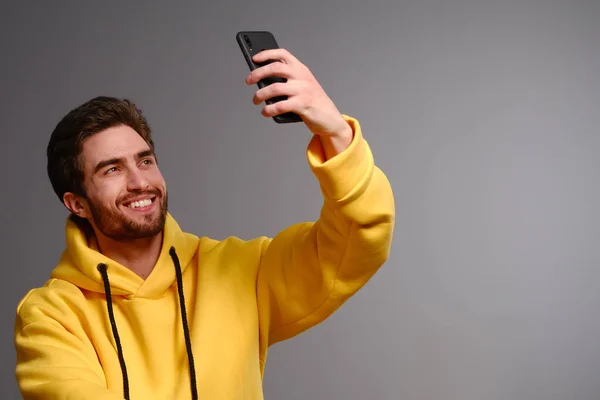 Joven chico sonriente en una sudadera amarilla con un vaso de papel desechable de café en la mano hace selfie en un teléfono inteligente —  Fotos de Stock