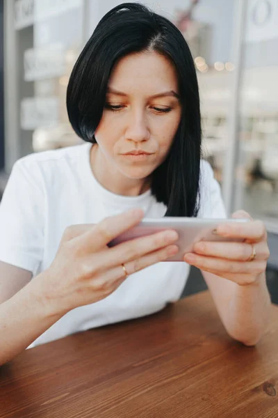 一位黑头发、身穿白色T恤的年轻女子坐在咖啡店的露天阳台上，一边用智能手机等候点菜，一边走人. — 图库照片