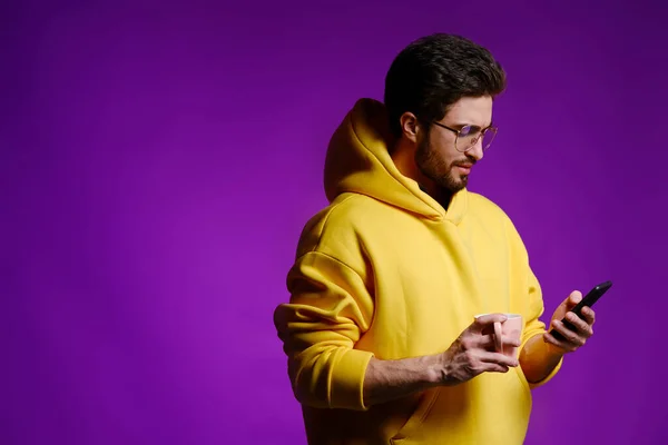 Молодой человек 25-30 лет в очках и в желтой толстовке с розовой кружкой в одной руке и смартфоном в другой на фиолетовом фоне . — стоковое фото