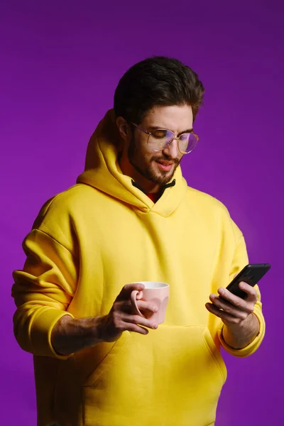Młody mężczyzna 25-30 lat w okularach i żółta bluza trzymający różowy kubek w jednej ręce i smartfon w drugiej na fioletowym tle. — Zdjęcie stockowe