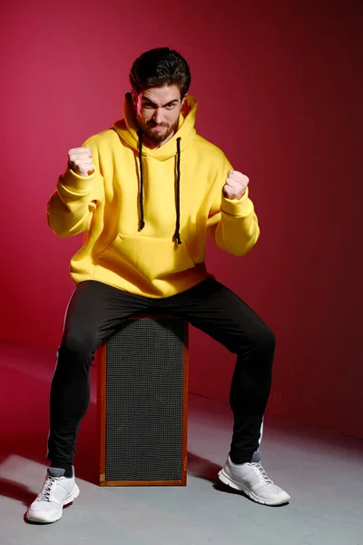 Un jeune homme de 25-30 ans en sweat-shirt jaune assis sur une vieille colonne de musique sur fond de mur rose et montrant différentes émotions. — Photo