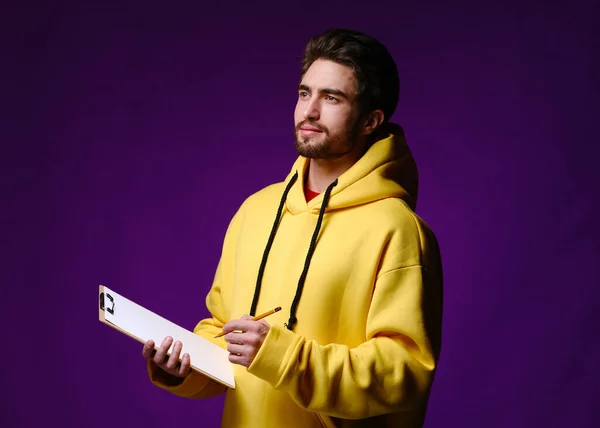 A young man of 25-30 years in glasses and a yellow sweatshirt holding a clipboard with clip in one hand and a pencil in the other — Stock Photo, Image